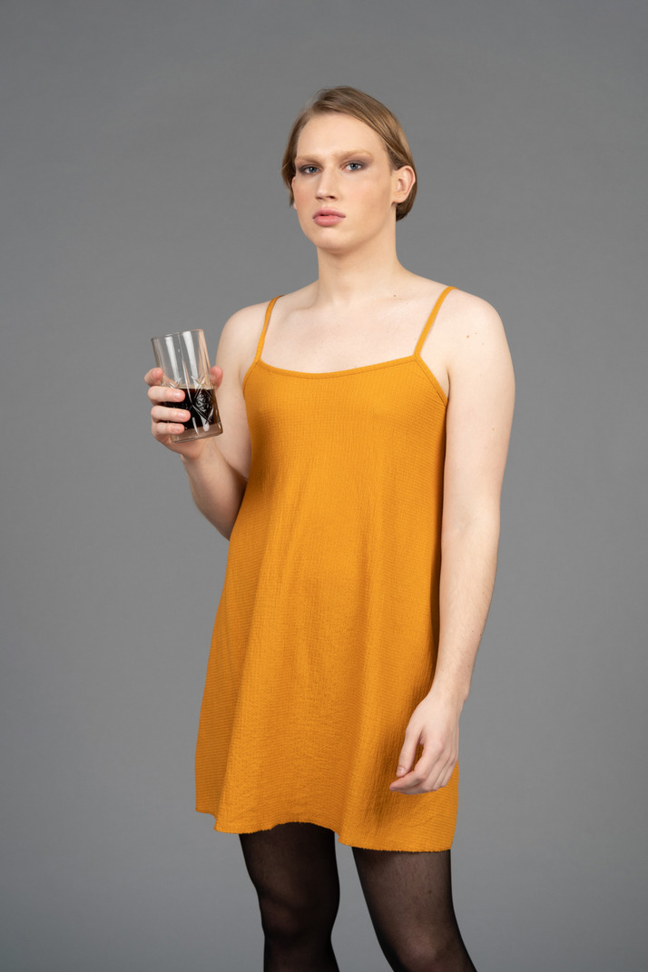 Young genderqueer person in orange dress looking pensive with drink in hand