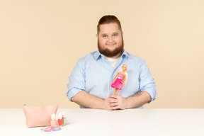 Sonriente tímido joven grande sentado a la mesa y sosteniendo la muñeca barbie