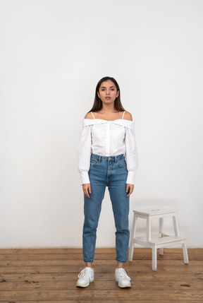 A young woman in white blouse standing next to a white chair in front of a white wall