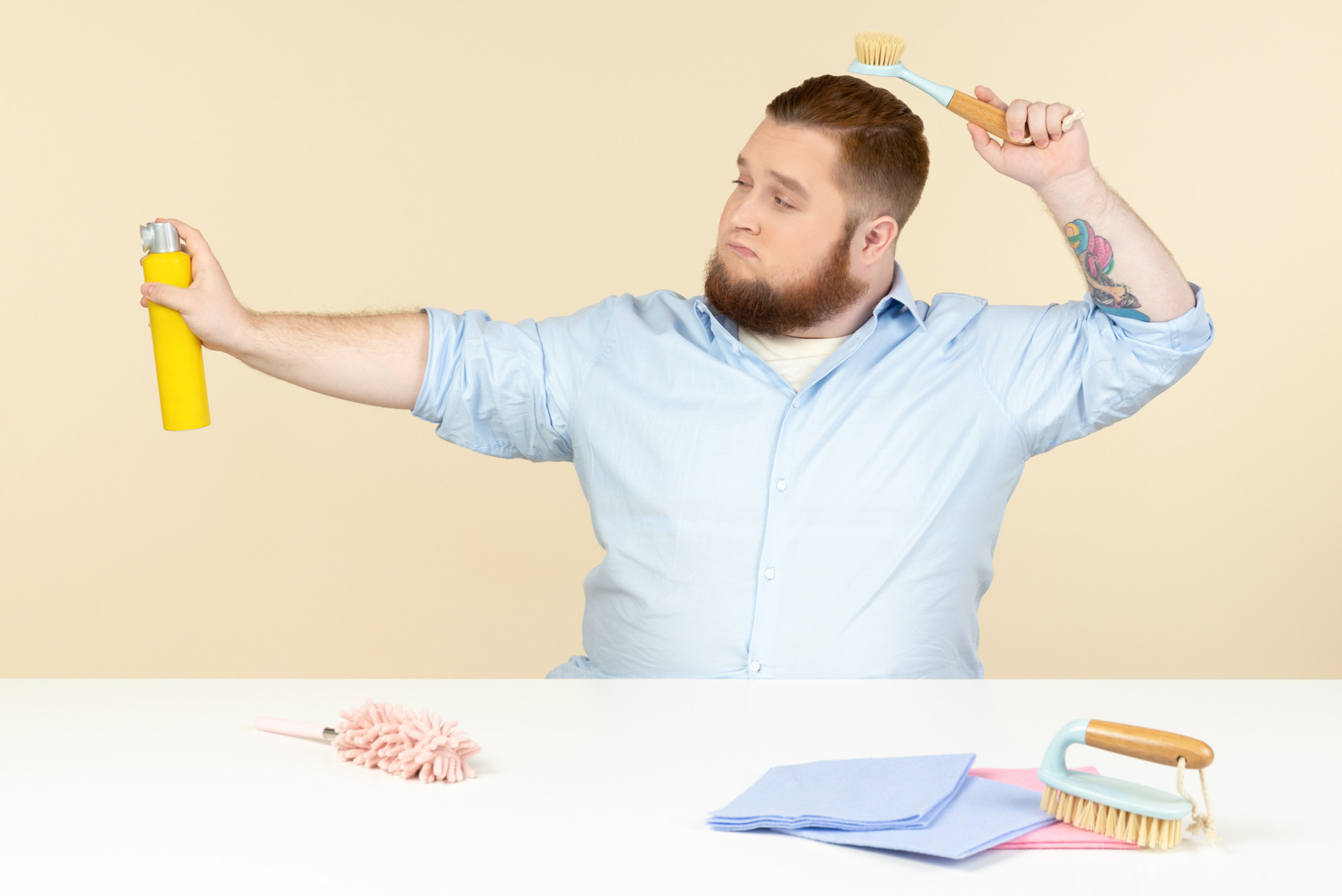 Young overweight man sitting at the table in the profile and holding cleaning tools
