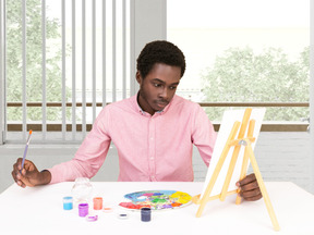 A man sitting at a table and painting on canvas with paint palette