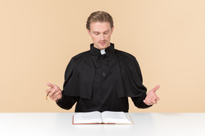 Catholic priest sitting at the table with open bible book and like saying something