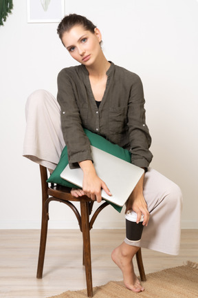 Front view of a young woman sitting on a chair and holding her laptop & touching coffee cup