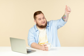 年轻的超重男子坐在笔记本电脑前的桌子和喝茶