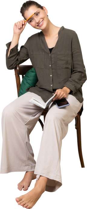 Front view of a smiling young woman wearing home clothes sitting on a chair with pencil and notebook