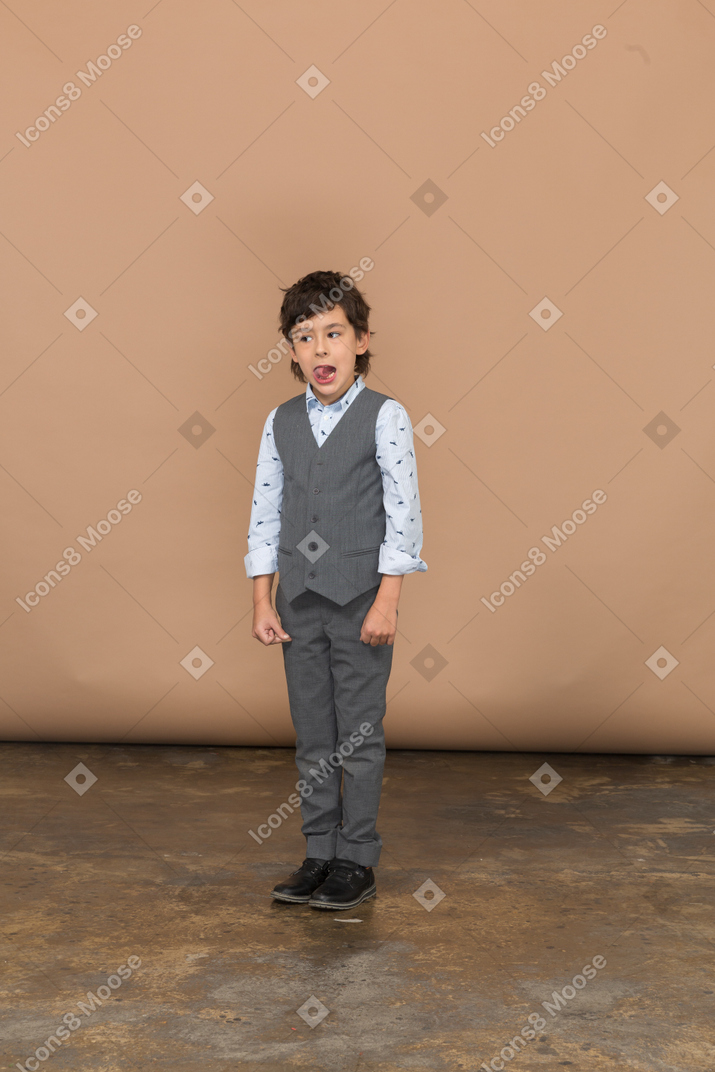 Vista frontal de um menino fofo em um terno cinza olhando para o lado e mostrando a língua