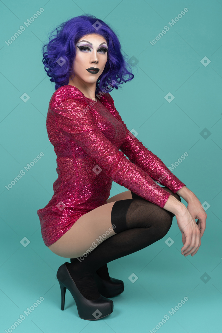 Side view of a drag queen in pink sequin dress squatting with hands on knees