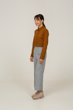 Vista di tre quarti di una giovane donna asiatica sorridente in calzoni e camicetta in piedi ancora
