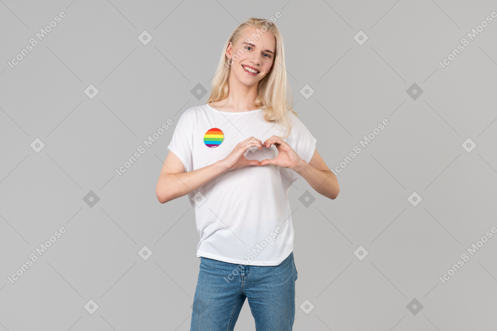 Good-looking young man with long blond hair, standing against grey background, wearing blue jeans and a white t-shirt with lgbt badge on it