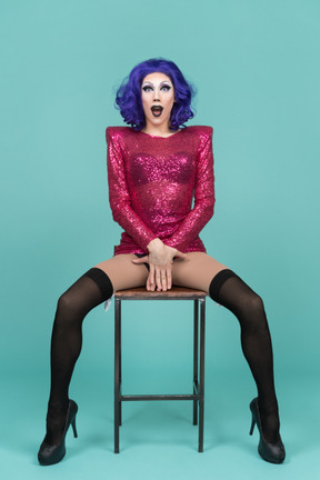 Drag queen covering privates with hands while sitting on a stool