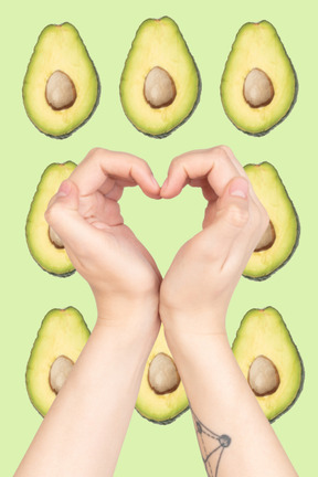 A person making a heart shape with their hands with avocados in the background