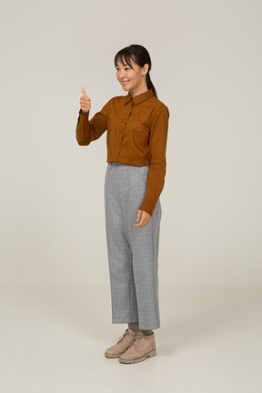Vista di tre quarti di una giovane donna asiatica in calzoni e camicetta che mostra il pollice in su