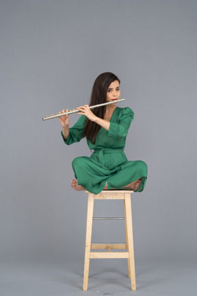 Toute la longueur d'une jeune femme jouant de la clarinette assise, les jambes croisées sur une chaise en bois