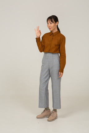 Vista laterale di una giovane donna asiatica in calzoni e camicetta che mostra gesto ok