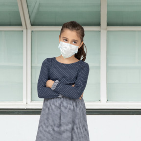 A young girl wearing a face mask standing in front of a window