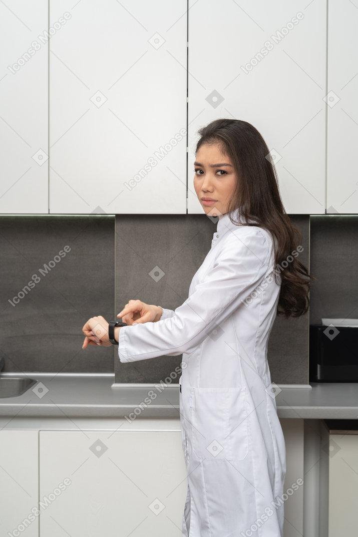 Frowning woman in white coat pointing at smartwatch