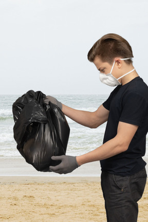 A man wearing a face mask and gloves holding a bag of garbage on a beach
