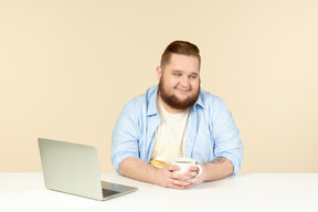 Довольный молодой грузный сидя перед ноутбуком и пьет чай