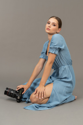 Side view of a young woman in blue dress sitting on a floor with camera