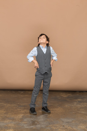 Vista frontal de um menino de terno cinza posando com a mão no quadril e olhando para cima