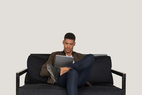 Front view of a young man sitting on a sofa while holding tablet