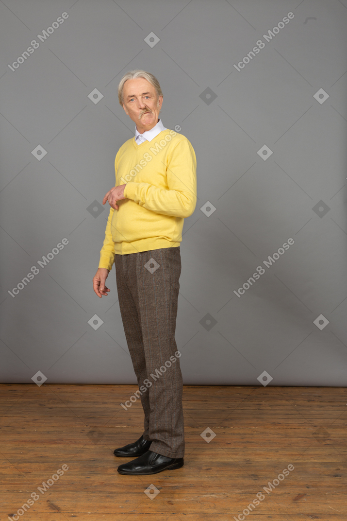Vista de três quartos de um velho suspeito em um pulôver amarelo, levantando a mão e olhando para a câmera