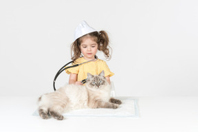 Little kid girl with stethoscope and wearing medical hat curing a cat