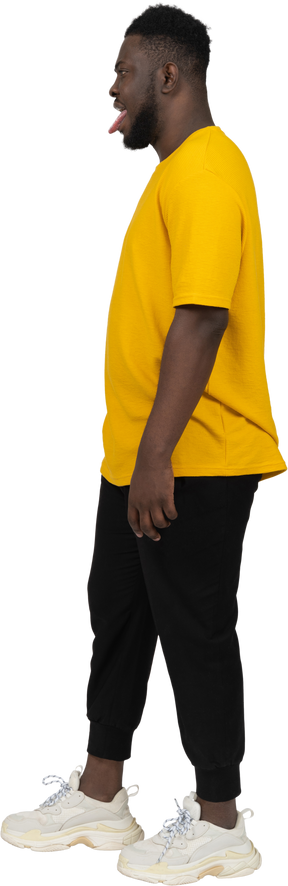 Side view of a young dark-skinned man in yellow t-shirt standing still & showing tongue