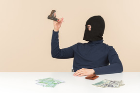 Hacker in balaclava sitting at the table with money bills on it and playing with a gun