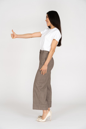 Vista lateral de uma jovem sorridente de calça e camiseta mostrando o polegar