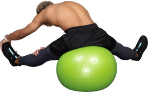 Vista posteriore di un uomo afro a torso nudo che si allunga mentre è seduto su una palla da palestra verde