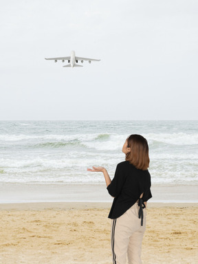 A woman standing on a beach looking at an airplane in the sky