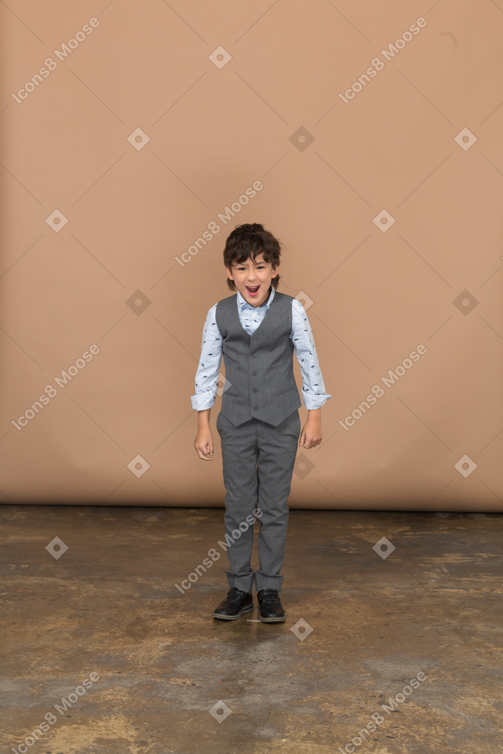 Vista frontal de un niño enojado en traje de pie con los puños cerrados y mirando a la cámara