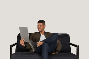 Vista frontal de um jovem sorridente sentado em um sofá enquanto assiste ao tablet