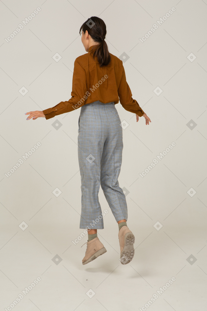 Vista posteriore di tre quarti di una giovane donna asiatica che salta in calzoni e camicetta allargando le mani