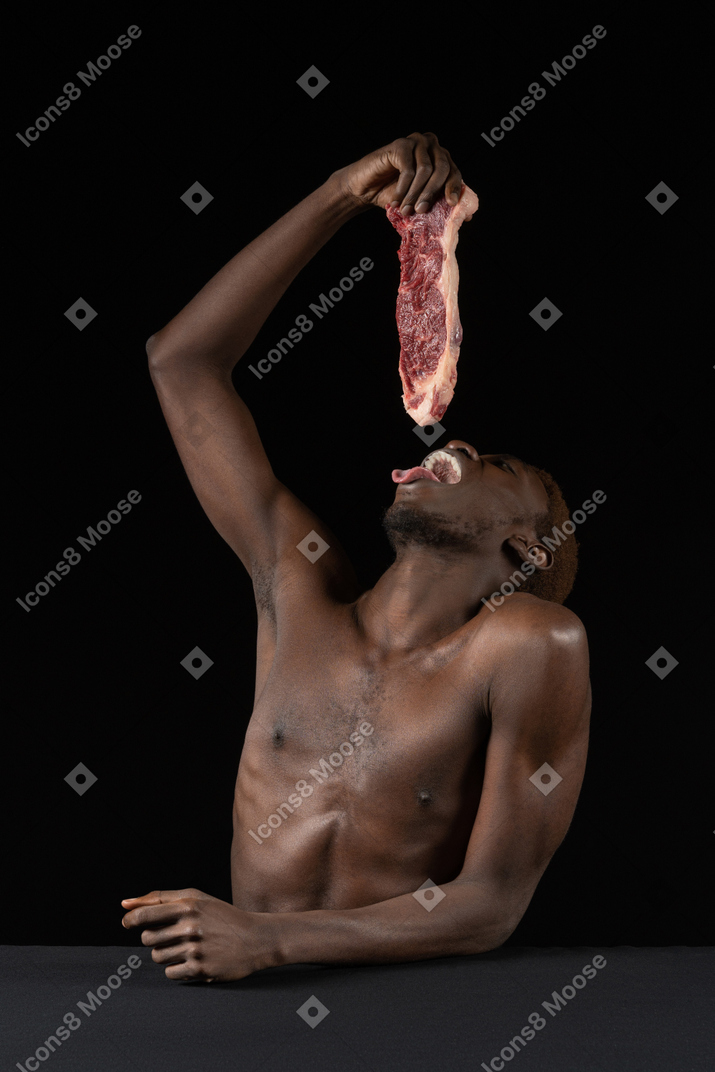 Front view of a young afro man holding a slice of meat while opening mouth wide