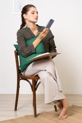 Vue de face d'une jeune femme réfléchie assise sur une chaise avec une tablette tout en vérifiant son téléphone