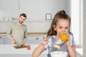 A man cutting vegetables with a little girl eating cereals in the kitchen