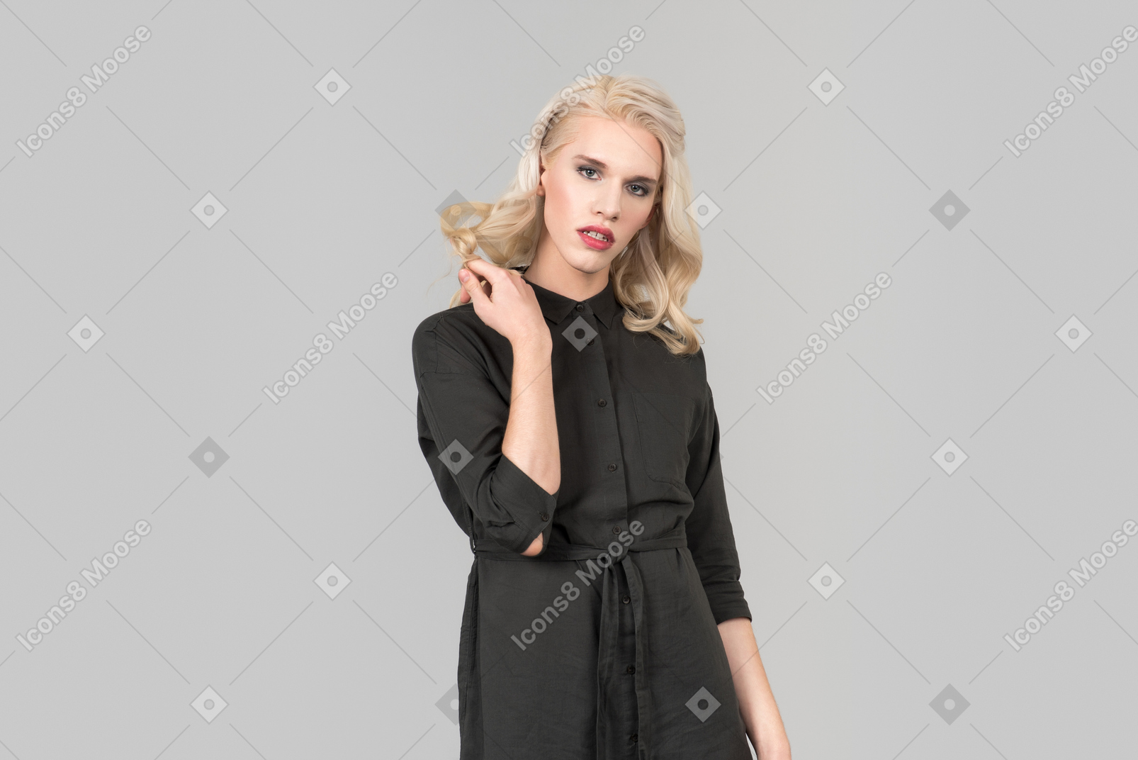 A young blond-haired person in a black dress  standing against the plain grey background