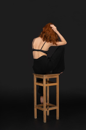 A three-quarter view of the beautiful woman dressed in black pants and bra, sitting on the wooden chair and holding her hand on the hair