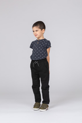 Вид спереди симпатичного мальчика в повседневной одежде, стоящего с руками за спиной и гримасничающего