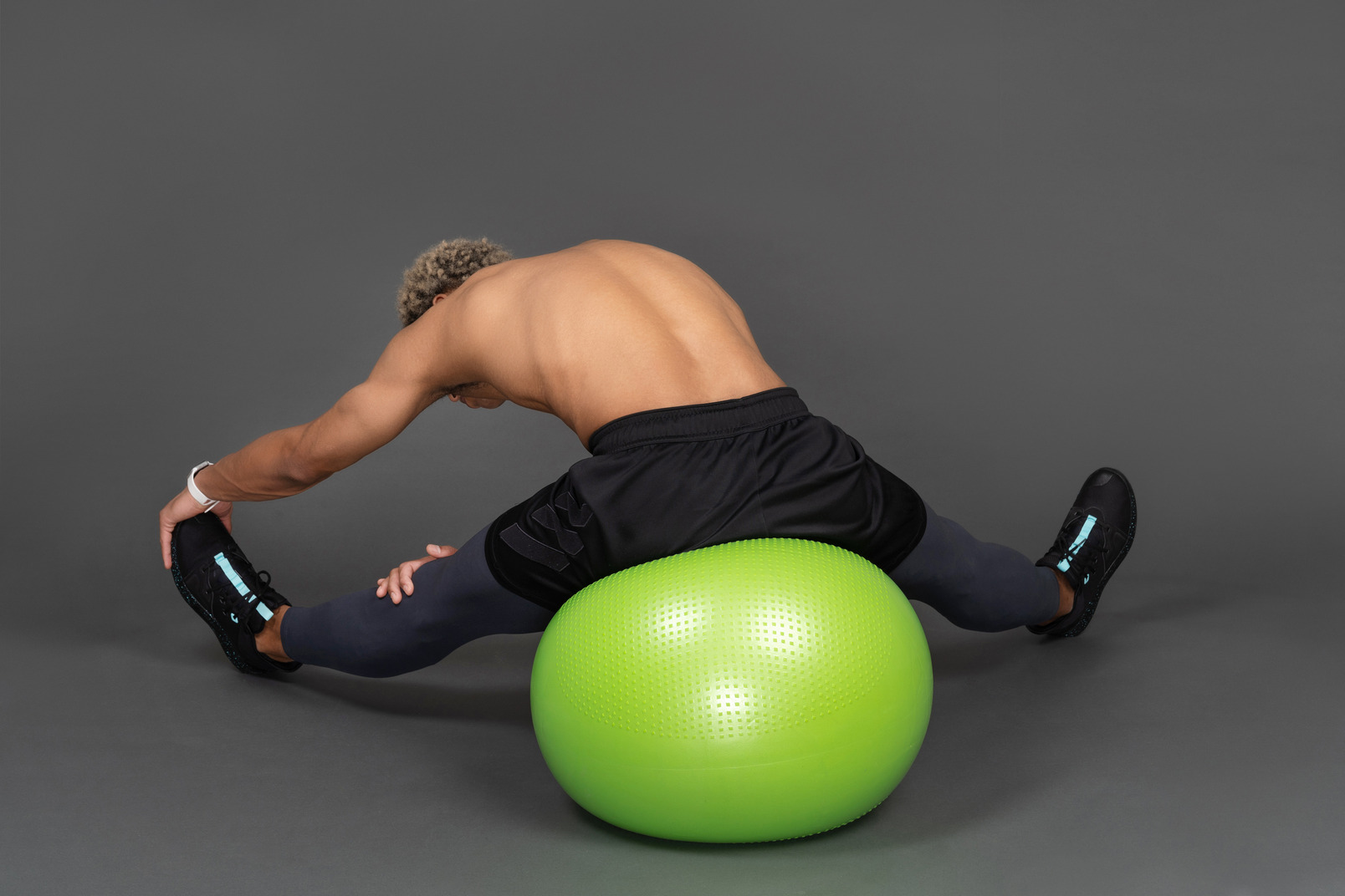 Back view of a shirtless afro man stretching while sitting on a green gym ball