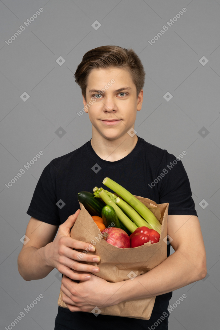 Junger mann, der kamera mit einer papiertasche betrachtet, die mit früchten gefüllt ist