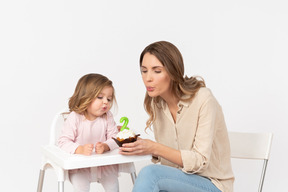 Menina soprando uma vela como a mãe dela está segurando um bolo de aniversário