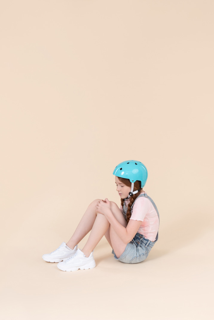 Teenager girl in blue helmet sitting on the floor and seems like got her knee hurt