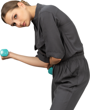 Вид сбоку молодой женщины в комбинезоне, делающей упражнения с гантелями