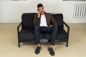 Front view of a perplexed young man sitting on a sofa and talking on his phone