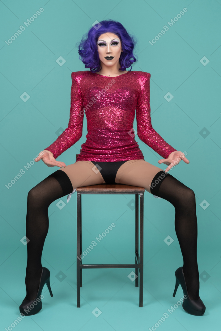 Drag queen sentada com as pernas abertas e apontando para a virilha