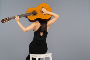 Back view of a young lady in black suit holding the guitar behind head and sitting on stool