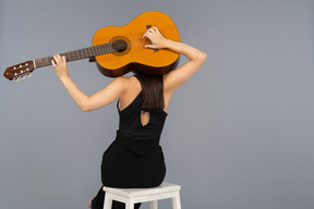 Vista posterior de una joven en traje negro sosteniendo la guitarra detrás de la cabeza y sentada en un taburete